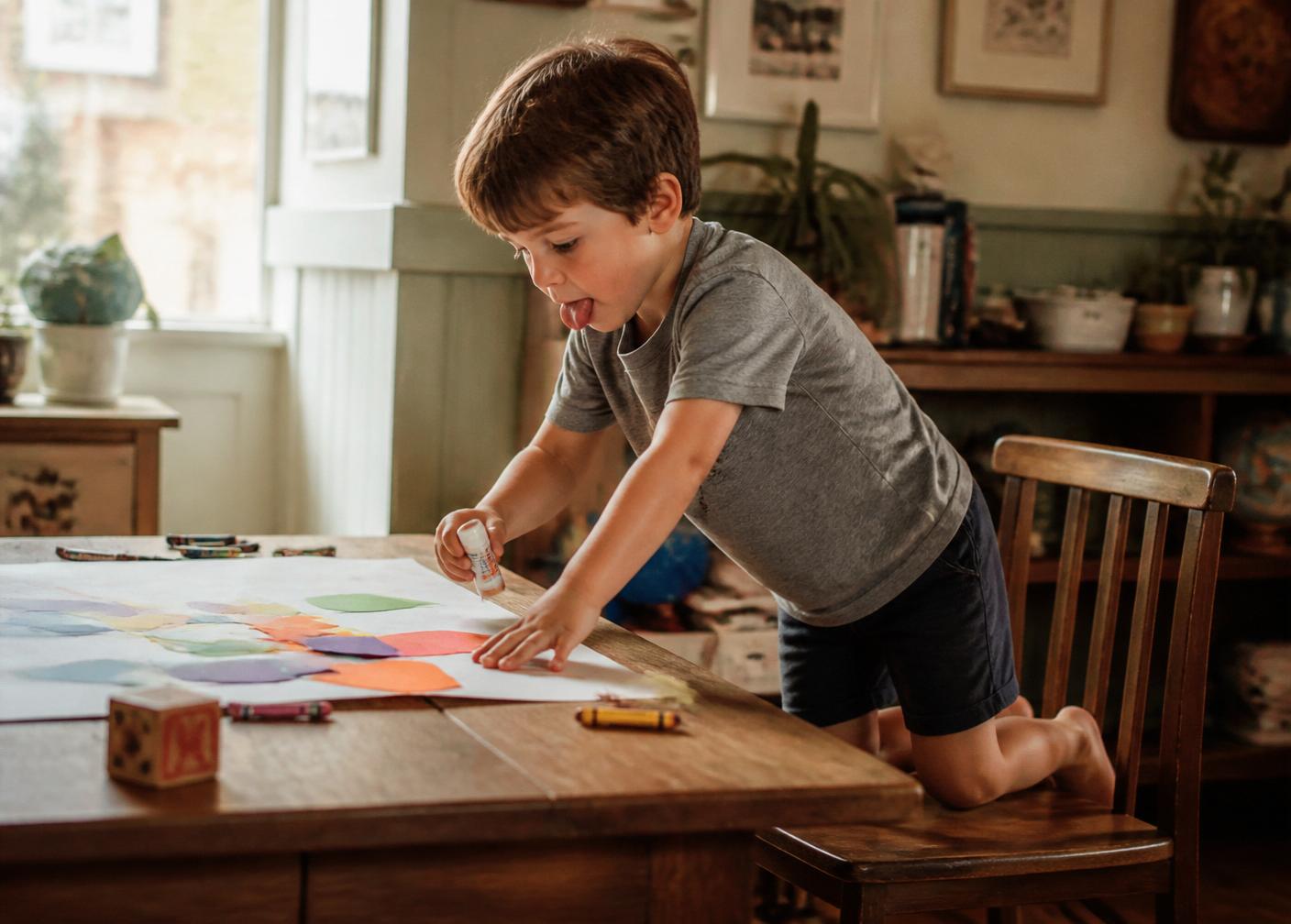 A young boy kneeling on a chair, gluing paper shapes onto a large sheet